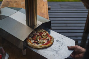 Man is pulling a delicious fresh home made pizza out of a stainless steel home portble oven fueled by pellets. Outdoor pizza party.