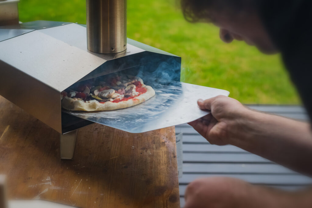 Man is pulling a delicious fresh home made pizza out of a stainless steel home portable oven fueled by pellets. Outdoor pizza party.