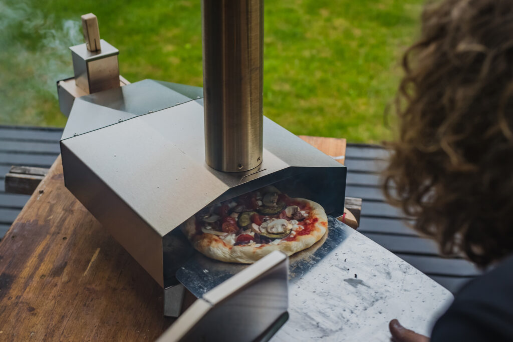 Man is pulling a delicious fresh home made pizza out of a stainless steel home portble oven fueled by pellets