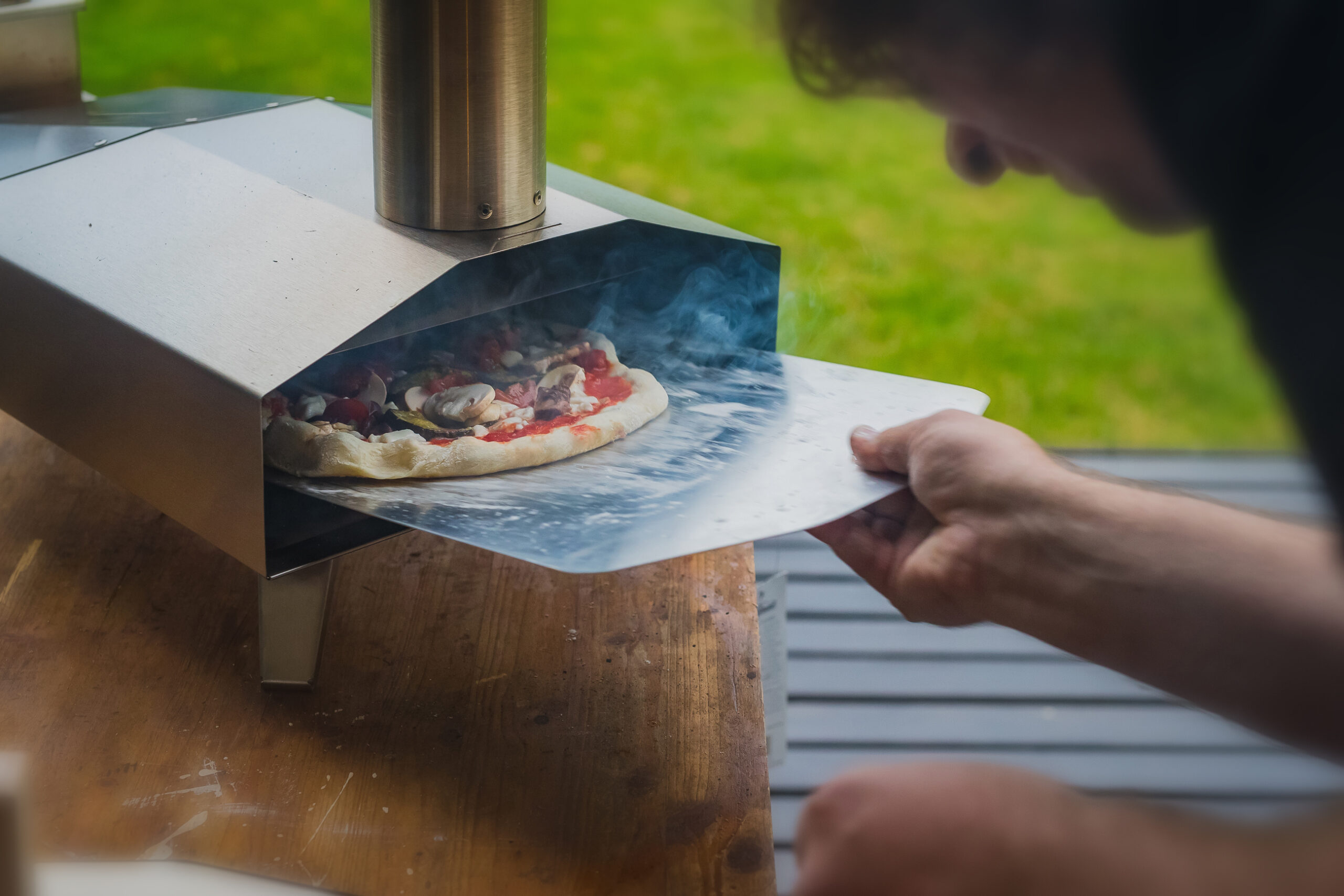 Man is pulling a delicious fresh home made pizza out of a stainless steel home portble oven fueled by pellets. Outdoor pizza party.