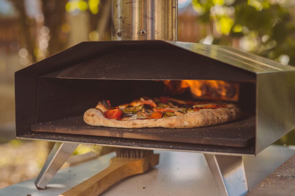 Home made pizza is inserted in a portable aluminium home oven for pizzas. Delicious pizza is baking in an oven, visible fire in the back of the furnace.